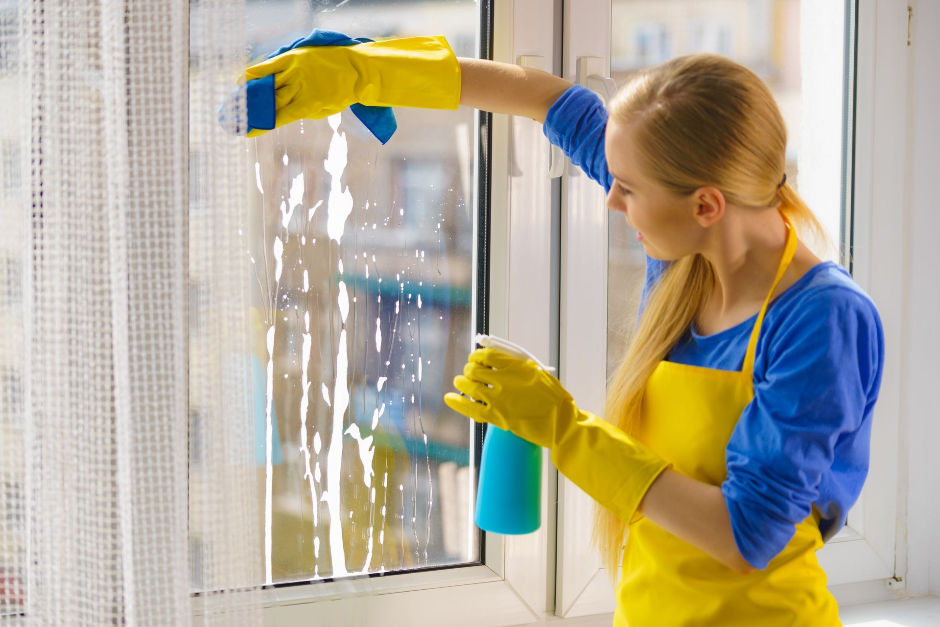 Young woman in yellow gloves cleaning window pane at home with rag and spray detergent. Cleaning concept